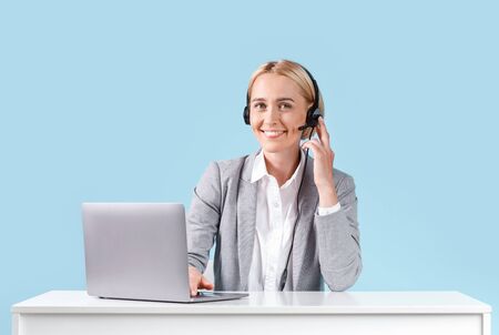 Beautiful female helpdesk operator with laptop and headset sitting at table against blue background