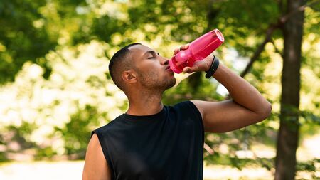 Stay hydrated. Sporty black guy drinking water from plastic bottle after his training at park. Panoramaの写真素材