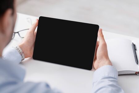 Video conference. Man in light shirt at table holds tablet with blank screen, glasses, notebook and pen nearの写真素材