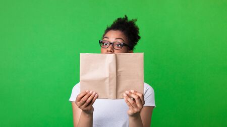 Bookworm Concept. Attentive african female nerd in eyeglasses holding and reading book on green studio wall, copy spaceの写真素材