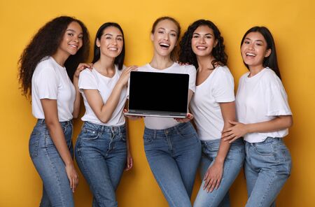 Five multiethnic women holding laptop showing blank computer screen recommending girlish website standing over yellow studio background. Mockupの写真素材
