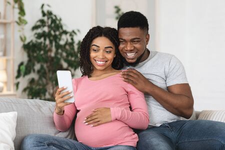 Happy black expecting couple taking selfie while resting at home, using smartphoneの写真素材