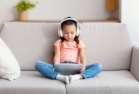 Gadget Addiction. Chinese Girl Using Digital Tablet And Wireless Earphones Watching Cartoons Sitting On Sofa Indoorの写真素材