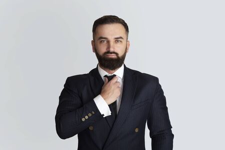 Serious businessman in office wear touching his necktie on grey background