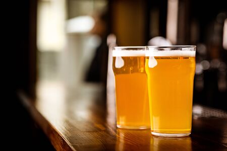 Evening meeting in pub. Two glasses with light beer on wooden bar counter in interior, on blurred...