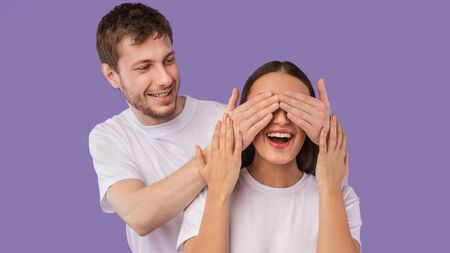 Guess Who. Playful young boyfriend covering his girlfriends eyes from behind, surprising her, pastel...