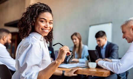 African American Business Lady Sitting On Corporate Meeting With Partners Smiling To Camera In Modern Office. Empty Spaceの写真素材