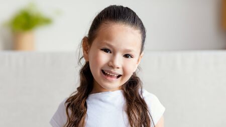 Adorable Asian Kid Girl Smiling To Camera Posing Sitting On Couch At Home. Little Girls Portrait....