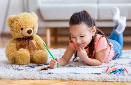 Asian Kid Girl Drawing Picture Using Crayon Lying On Floor At Home. Little Artist, Childs Hobby...