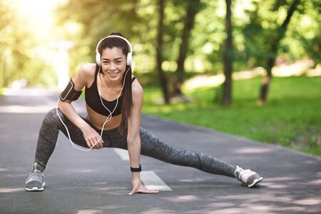 Sporty Asian Woman In Headphones Warming Up Before Jogging In Park, Stretching Legs And Smiling At...