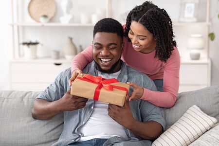 Gift With Love. Caring Black Wife Surprising Happy Husband With Romantic Present At Home, Greeting With Anniversary Or Birthdayの写真素材