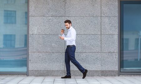 Confident CEO with coffee and mobile phone walking near office building in city center, panoramaの写真素材