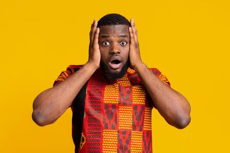 Shocked african guy in traditional costume with open mouth holding his head, yellow studio...