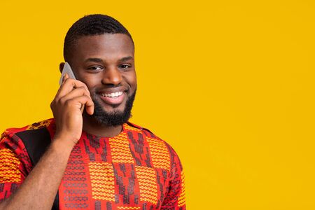 Closeup of smiling black man in traditional african clothes talking on mobile phone over yellow...