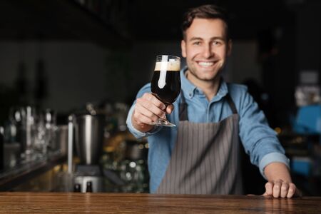 Cheers. Smiling barman in apron holds out glass of dark beer with foam, for client in pub interior, free spaceの写真素材