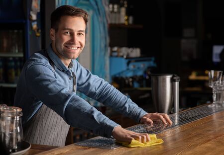 Welcome to bar. Smiling handsome barman with rag in his hands, behind the rack in interior of pubの写真素材