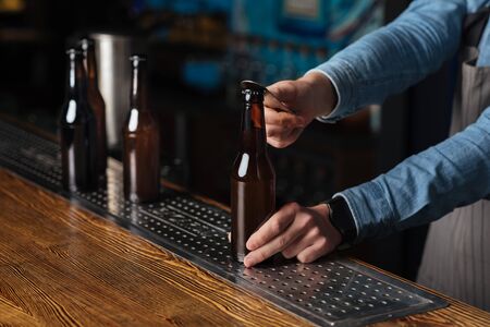 Cold drink for clients. Barman open bottle of beer on wooden bar in interior, cropped