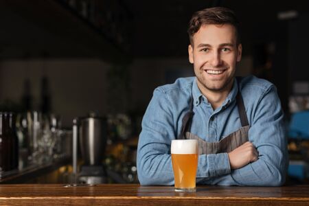 Beer traditions in modern pub. Smiling handsome guy in denim shirt leans on bar counter with glass of light ale with foam, in interior, free spaceの写真素材