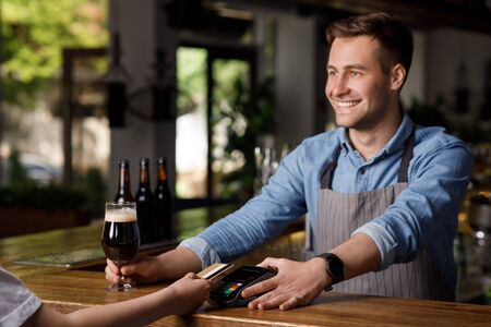 Modern payment service in pub. Smiling barman with terminal and glass of dark beer, looks at...