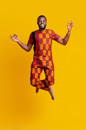 Carefree african guy in bright costume jumping up over yellow studio backgroundの写真素材