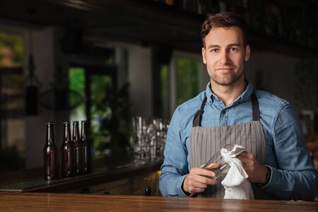 Modern work of bartender. Handsome man in apron with stylish haircut, rubs glass, standing at bar counter in interior pubの写真素材