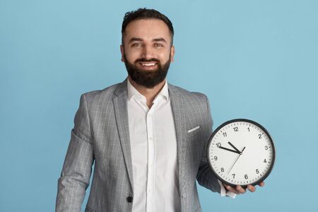 Time management and optimization concept. Happy businessman holding analog clock on blue background