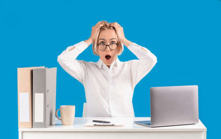 Mature secretary holding her head in shock at her workplace with laptop computer, blue studio...