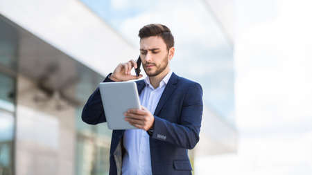 Serious company director with tablet computer talking on smartphone on city street, panoramaの写真素材