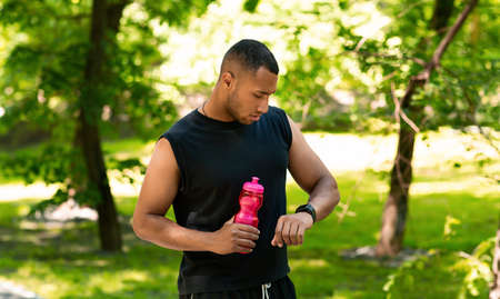 African American runner with bottle of water checking his smartwatch after workout at park, panorama