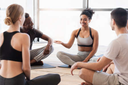 Black woman yoga instructor talking to young yogi group after class, sitting on floor in studio and chatting togetherの写真素材