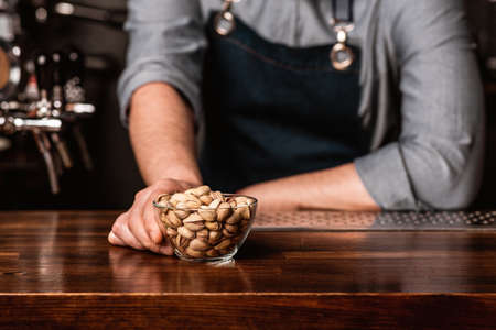 Order snack at pub. Barman in apron gives to client glass plate with pistachios on wooden bar counter, cropped, free spaceの写真素材
