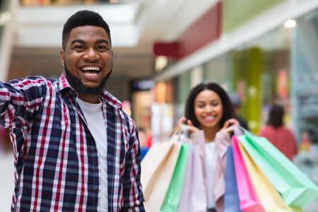 Cheerful black couple taking selfie on cellphone in department store, having fun during shopping, selective focus on handsome guyの写真素材