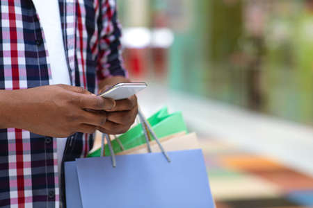 Cashback Concept. Unrecognizable Black Man With Shopper Bags Using Smartphone In Mall After Shopping, Checking Money Bonus On Account, Croppedの写真素材