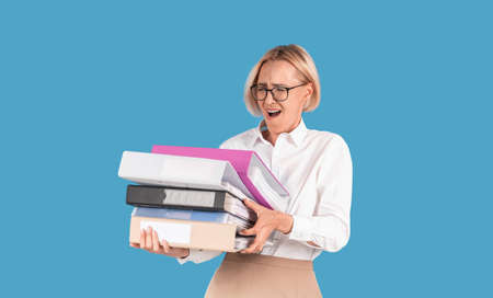 Stressed female secretary overloaded with paperwork, holding folders with documents on blue studio...