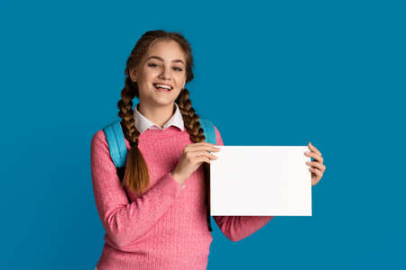 Back to school. Smiling girl school girl holds white sheet with empty space isolated on blue background, studio shotの写真素材