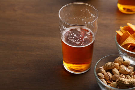 Beer party. Half glass of light ale in glass stands on wooden table, near pistachios and nachos in bowlsの写真素材