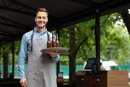 Work a waiter. Smiling handsome man in apron holds tray with bottles of beer in hands on open area of pub in daytimeの写真素材