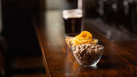 Menu for client. Pistachios, chips, nuts and glass of dark beer with foam on bar counter in interior of pub, close up, panorama, free spaceの写真素材