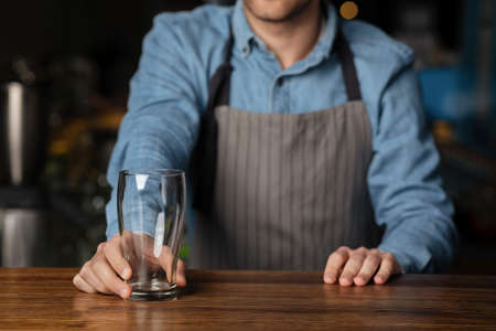 Customer service in pub. Barman in apron gives empty glass to client on wooden bar counter in...