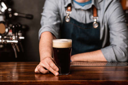 Dark beer with foam in glass from bartender. Guy in apron gives drink on wooden counter in interior...