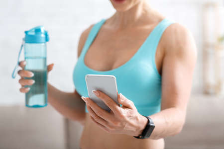 Healthy lifestyle and sport at home. Muscular woman in sportswear with smart watch holds bottle of water and smartphone in hands, cropped, close up, free spaceの写真素材
