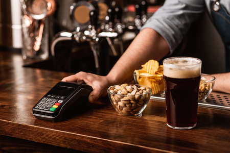 Payment for beer set. Bartender gives contactless terminal, near pistachios, chips and nuts in plates, and dark beer with foam in interior of pubの写真素材