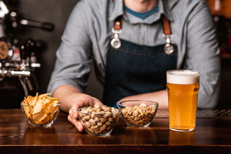 Crispy snacks from the bartender. Pistachios, nuts and chips in plates and glass of light beer with foam on wooden bar counter, croppedの写真素材