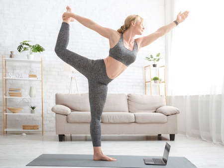 Yoga practice and online home lessons on quarantine. Adult woman doing exercise, stands on mat with laptop in living room interior, copy spaceの写真素材