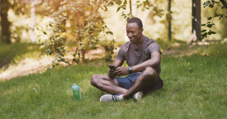 Rest in park. African american guy using smartphone, sitting on grass outdoors, free space