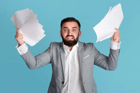 Dealing with workplace stress. Angry overworked office employee grasping paper documents on blue backgroundの写真素材