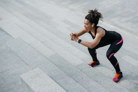 Jumping and squats on stairs. Smiling african american girl with wireless headphones and fitness tracker doing exercises on stairs in city, free spaceの写真素材