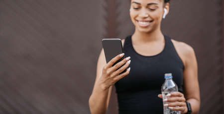 Sports and biorhythms. Happy african american girl with headphones and fitness tracker holds bottle...
