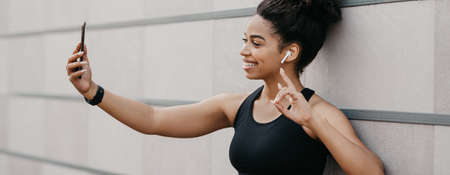 Selfie for sports blog. Muscular smiling african american girl in sport bra makes sign the peace and...