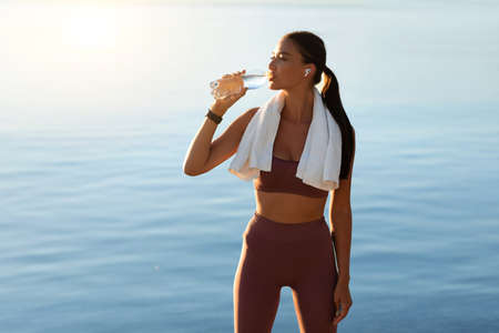 Wellness Concept. Active girl drinking mineral water from bottle, standing on the beach in wireless...
